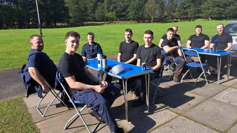 The Navy sit on metal chairs at two tables, with drinks, having a rest outside at Catterburn campsire