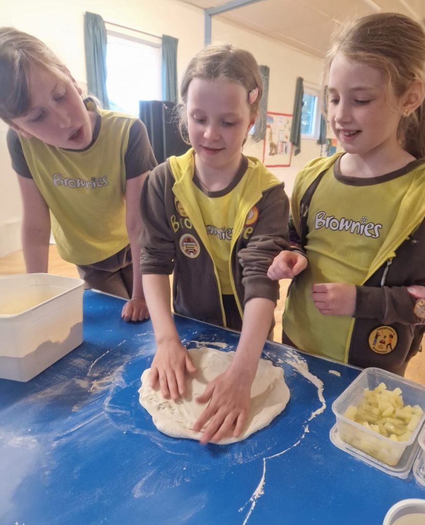 Three Brownies stand over a table, while one Brownie in the centre rolls out her pizza dough.