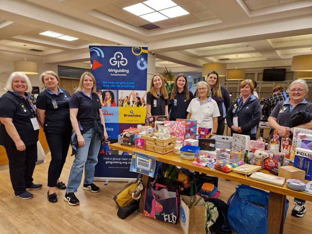 Members of Girlguiding Dunbartonshire's county team standing inside the hotel around a table filled with prizes. There's a tall Girlguiding banner stand as well